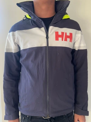 Helly Hansen vindjacka med huva blå, storlek S - Mörkblå vindjacka från Helly Hansen med vit bröstpanel och röd HH-logga. Jackan har en gul huva, hög krage och hel dragkedja framtill. Perfekt för blåsiga dagar och har sportig look. Lite slitningar på reflexer samt solblekt.