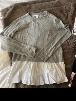 Grå tröja med vit skjortdetalj H&M - Snygg grå tröja från H&M i storlek M med vit skjortdetalj nedtill. Tröjan har rund halsringning, långa ärmar och ribbade muddar. Perfekt för en lager-på-lager-look och enkel att styla till jeans eller kjol.