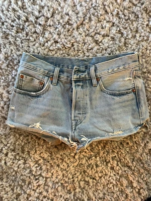 Levi's 501 ljusblå jeansshorts - Säljer ett par klassiska Levi's 501 jeansshorts i ljusblå denim. Shortsen har råa, fransiga kanter och slitna detaljer för en avslappnad look. Femficksmodell med bälteshällor och knappgylf. Perfekta för sommaren!