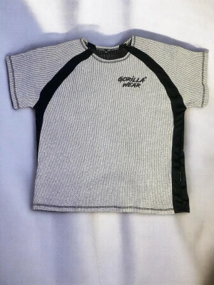  Gorilla Wear t-shirt - Snygg t-shirt från Gorilla Wear med svart och vit smalrandigt mönster och svarta paneler längs sidorna och axlarna. Tryckt logga på bröstet och ryggen. Klassisk passform och rund hals. Perfekt för dig som gillar sportig stil.