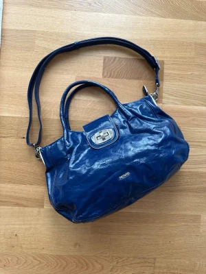 Blå handväska från Picard i skinn - Snygg blå handväska från Picard i glansigt skinn. Väskan har både handtag och justerbar axelrem, silverfärgade metalldetaljer och ett dekorativt lås framtill. Praktisk storlek med ytterfack och nyckelring med Picard-logga.