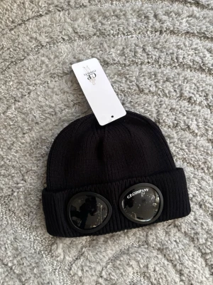 Svart mössa med glasögondetalj C.P. Company - Säljer en svart ribbstickad mössa från C.P. Company med ikoniska glasögonlinser på uppviket. Mössan är tillverkad i mjukt material och har en modern, streetinspirerad look. Perfekt accessoar för dig som gillar unik stil. 2st tillgängliga 