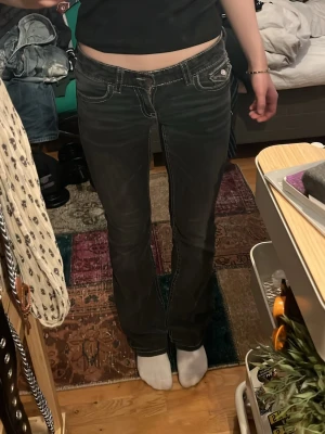 Svarta bootcut jeans med vita sömmar - Säljer ett par svarta bootcut jeans med vita kontrastsömmar och låg midja. Jeansen har fem fickor och en liten dekorativ knappdetalj på fickan fram. Snyggt slitna detaljer och klassisk passform som ger en retrovibe.