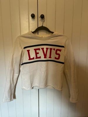  tröja från Levi's - Vit sweatshirt från Levi's med röd logga framtill och blåa linjer ovanför och under texten. Tröjan har rund halsringning och långa ärmar. Perfekt för en avslappnad och trendig stil.