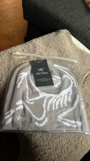 Grej mössa från Arc'teryx - Snygg ljuslila mössa från Arc'teryx med vit logga och grafiskt mönster på framsidan. Tillverkad i mjukt, stickat material som sitter skönt på huvudet. Perfekt för dig som vill ha en stilren och modern accessoar till höst och vinter.