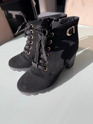 Svarta snörade stövletter med klack - Säljer ett par svarta stövletter med snörning och grov klack. Skorna har guldfärgade detaljer vid snörningen och ett spänne på sidan. Materialet ser ut att vara mockaimitation och de har dragkedja på insidan för enkel påtagning.