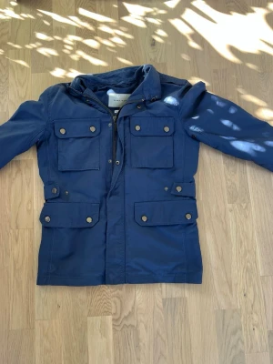 Marinblå vindjacka med fickor - Snygg marinblå field jacket perfekt till våren i storlek xs. Säljer pga för liten. Väldigt bra skick inga större tecken på användning. 