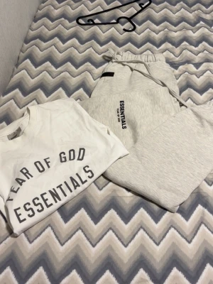 Grå tracksuit  från Fear of God Essentials - Snygg ljusgrå hoodie från Fear of God Essentials med svart logga på bröstet. Klassisk passform med huva och ribbade muddar. Tillverkad i mjukt bomullsmaterial som är perfekt för en avslappnad streetwear-look.