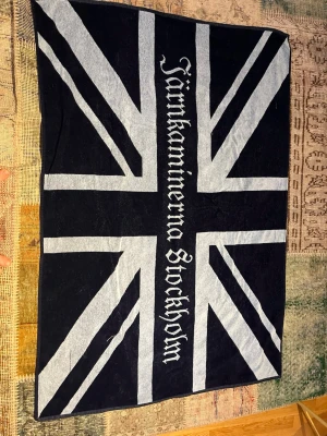 Strandhandduk Jänkarninerna Stockholm - Snygg strandhandduk,mönster i mörkblått och ljusblått samt texten 'Järnkaminerna Stockholm' i gotisk stil. Handduken är tillverkad i mjukt frottématerial och passar perfekt för stranden eller poolen.