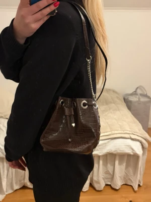 Brun axelväska i crocopräglat skinn - Snygg brun axelväska i crocopräglat konstläder med silverfärgade detaljer och kedja. Väskan har dragskostängning och justerbar axelrem. Perfekt storlek för det viktigaste och ger en trendig touch till din outfit.