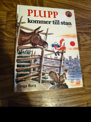 Plupp kommer till stan bok - En illustrerad barnbok med titeln 'Plupp kommer till stan' av Inga Borg. Omslaget är färgglatt med motiv av Plupp, en älg och en ekorre vid ett staket, samt en stad i bakgrunden. Boken har hårda pärmar och tydlig text på framsidan.