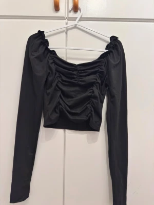 Svart croppad topp med rynk - Trendig svart croppad topp med långa ärmar och snygga rynkdetaljer framtill. Toppen har en offshoulder-design och är tillverkad i mjukt material som sitter bekvämt mot kroppen. Perfekt för dig som vill ha en stilren men ändå unik look.