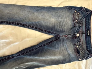 True Religion blå jeans med röd söm - Snygga blå jeans från True Religion med tydliga röda kontrastsömmar och klassisk femficksmodell. Jeansen har en tvättad look och silverfärgade knappar. Perfekta för dig som vill ha en cool och avslappnad stil.