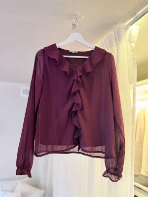 Plommonlila blus med volang - Snygg plommonlila blus i transparent material med långa ärmar och volang längs den djupa v-ringningen. Blusen har resår vid ärmsluten och en luftig passform som ger en trendig look.