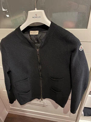 Svart Moncler cardigan  - Stilren svart ziptröja från Moncler med rund halsringning och två fickor framtill. Tröjan har ribbade muddar och Moncler-logga på ärmen. Perfekt för dig som gillar klassisk design och exklusiva material.