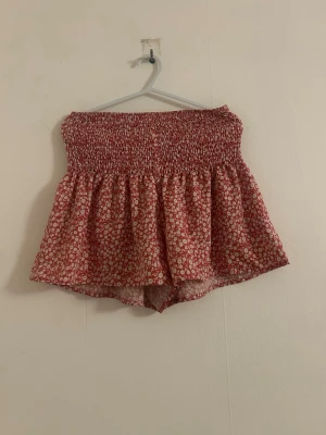Blommiga högmidjade shorts i rött - Säljer ett par söta högmidjade shorts med blommigt mönster i rött och vitt. Shortsen har en smockad midja som ger en bekväm och stretchig passform samt ett luftigt fall. Perfekta för varma dagar och enkel att matcha med olika toppar.