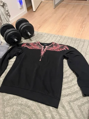 Svart sweatshirt med rosa vingtryck - Säljer en svart sweatshirt från Marcelo Burlon med ett fett rosa och svart vingmönster över axlar och bröst, som fortsätter på ryggen. Tröjan har rund hals och är gjord i mjukt bomullsmaterial. Perfekt för dig som vill sticka ut med en unik design.