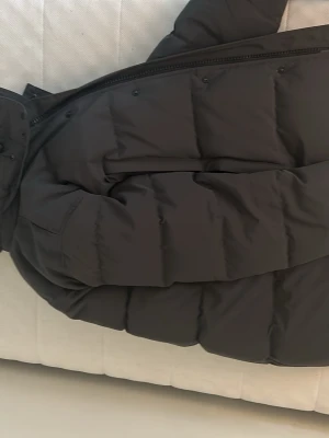 Grå dunjacka med pälskant black label - Säljer en grå dunjacka från Canada Goose med luva och pälskant. Jackan har en klassisk quiltad design, dragkedja och tryckknappar framtill samt en patch med logga på ärmen. Perfekt för kalla vinterdagar.