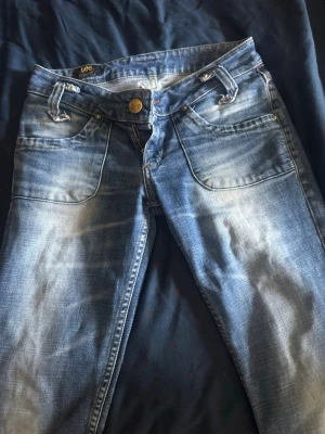 Blå Lee jeans med slitningar - Säljer ett par blå jeans från Lee med snygga slitningar och tvättade detaljer. Klassisk femficksmodell med guldknappar och Lee-logga på bakfickan. Jeansen är lätt bootcut och är gjorda i mjukt denimtyg. Perfekta för en avslappnad och cool stil. Älskar verkligen dessa men tyvär så är de lite för korta för mig. De är i längd 31 men är nedtagna men skulle säga att de passar nån som är cirka 165cm. En liten flärp är bortklippt på framsidan av jeansen men inget man tänker på. Nyskick! Strl 28