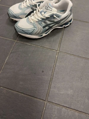 Vita och blå Asics sneakers - Snygga sneakers från Asics i vitt och ljusblått med svarta detaljer. Ovandelen är i mesh och syntet för bra andningsförmåga och komfort. Klassisk sportig look med lågt skaft och robust sula. Perfekta för dig som gillar retrostil och streetwear.