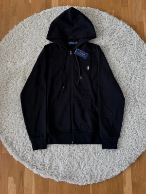 Svart Polo Ralph Lauren hoodie  - Snygg svart hoodie från Polo Ralph Lauren med dragkedja framtill, justerbar huva och klassisk logga broderad på bröstet. Hoodien har känguruficka och ribbade muddar vid ärmslut och nederkant. Perfekt för en avslappnad och stilren look. Pris kan diskuteras!