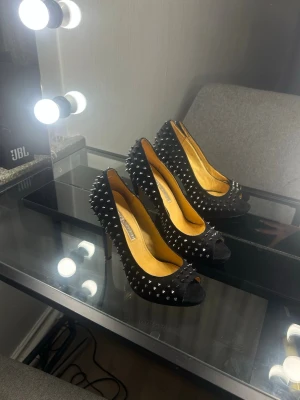 Svarta pumps med nitar från Buffalo London - Säljer ett par svarta pumps från Buffalo London med öppen tå och höga klackar. Skorna är täckta med silverfärgade nitar. Materialet är mockaimitation och insidan är orange. Perfekta för dig som vill sticka ut.