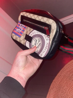 H&M axelväska med GG-mönster - Säljer en snygg axelväska från h&m med klassiskt GG-mönster, svart läderdetalj och färgglada tryck med texten 'AVEUGLE PAR AMOUR' och en tecknad figur. Väskan har en dragkedjeficka framtill och justerbar axelrem i grönt och rött.