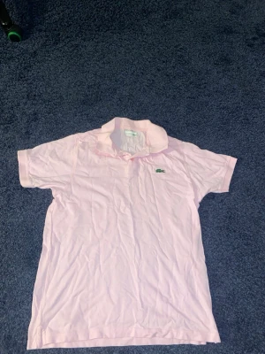 Ljusrosa pikétröja från Lacoste - Säljer en ljusrosa pikétröja från Lacoste med klassisk krage och korta ärmar. Tröjan har den ikoniska gröna krokodilloggan broderad på bröstet och är tillverkad i mjuk bomull. Perfekt för en avslappnad och stilren look.