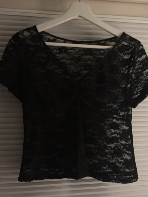 Svart spets topp med v-ringning - Säljer en svart topp i spets med v-ringning och korta ärmar. Toppen har ett snyggt rynkat parti framtill och är helt genomskinlig med blommigt spetsmönster. Perfekt för att skapa en cool och trendig look.