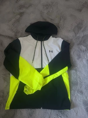 Under Armour vindjacka  - Snygg vindjacka från Under Armour i svart, vitt och neon-gult. Jackan har huva med dragsko, hel dragkedja framtill och elastiska muddar vid ärmsluten. Perfekt för dig som vill sticka ut med sportig stil. Tillverkad i lätt och tåligt material.