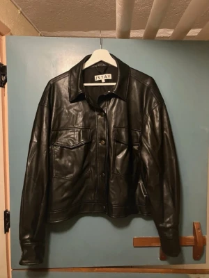 Svart skinnjacka från ISTAY - Snygg svart skinnjacka från ISTAY med klassisk krage och två stora bröstfickor med lock. Jackan har knäppning framtill och detaljer vid ärmslut och midja. Perfekt för dig som vill ha en stilren och tidlös look.