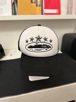 Svartvit truckerkeps med stjärnor - Säljer en svartvit truckerkeps med broderade stjärnor och ett UFO-motiv framtill. Kepsen har svart nät på sidorna och bak, samt böjd skärm. Justerbar storlek med snapback-spänne. Perfekt för dig som gillar streetwear och unika detaljer.