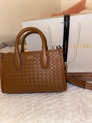 Brun handväska från BOSS - Snygg brun handväska från BOSS med flätad läderlook och guldfärgad logga. Väskan har både handtag och en bred axelrem med BOSS-tryck. Perfekt storlek för det viktigaste och ger en trendig touch till din outfit.