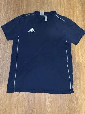 Mörkblå Adidas t-shirt med vita detaljer - Sportig mörkblå t-shirt från Adidas med vita kontrastsömmar och Adidas-logga på bröstet. Tillverkad i ett lätt och snabbtorkande material som passar perfekt till träning eller gym. Klassisk rund hals och korta ärmar.