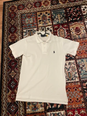Vit pikétröja från Ralph Lauren - Klassisk vit pikétröja från Ralph Lauren med den ikoniska broderade loggan på bröstet. Tröjan har korta ärmar, krage och knappar vid halsen. Perfekt för en clean och stilren look.
