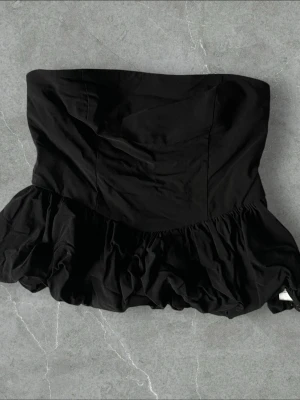 Svart axelbandslös volangtopp - Snygg svart axelbandslös topp med volangkant nedtill. Toppen har en bandeauformad överdel och smockad rygg för extra komfort. Perfekt för dig som vill ha en trendig och stilren look.