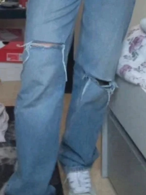 Ljusblå ripped jeans med raka ben - Säljer ett par ljusblå jeans med raka ben och stora slitningar vid knäna. Jeansen har klassisk femficksmodell och mid waist. Perfekta för en avslappnad och trendig look. Materialet är denim i bomull.