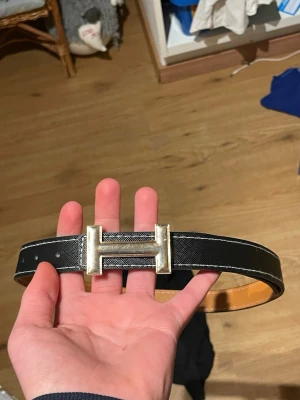 Svart bälte med H-spänne - Snyggt svart bälte med det ikoniska H-spännet i silverfärg. Bältet har vit kontrastsöm längs kanterna och är gjort i exklusivt skinn. Perfekt accessoar för att lyfta vilken outfit som helst.