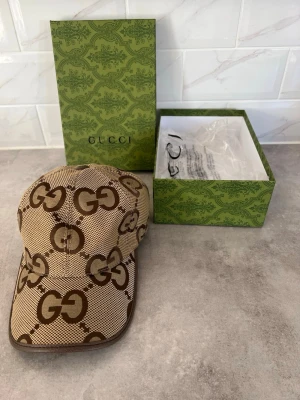 Gucci Keps - Säljer en beige keps från Gucci med det klassiska bruna GG-monogrammet över hela kepsen. Kepsen har böjd skärm, justerbar rem bak och är tillverkad i ett slitstarkt tyg. Kommer med originalkartong och tagg kvar.  Pris kan ev disskuteras, enbart seriösa köpare. Kan vid efterfrågan även mötas i Stockholm. Allt är i nyskick