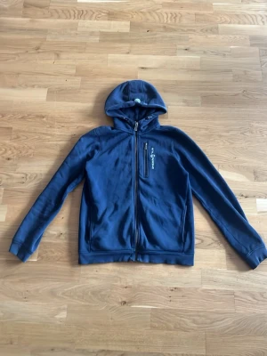 Blå hoodie från sail racing  - Mörkblå hoodie från sail racing  med dragkedja framtill och en bröstficka med logga. Tillverkad i mjukt material och har en klassisk huva. Perfekt för en avslappnad och sportig stil.