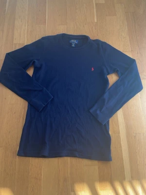 Mörkblå långärmad tröja Polo Ralph Lauren - Mörkblå långärmad tröja från Polo Ralph Lauren med rund hals och den klassiska röda logotypen broderad på bröstet. Tröjan är ribbad och har en slim fit passform som sitter snyggt på kroppen. Perfekt för en clean och stilren look. Perfekt för sommaren!