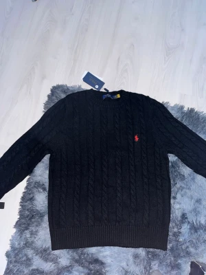 Svart kabelstickad tröja Polo Ralph Lauren - Kabelstickad svart tröja från Polo Ralph Lauren med rund halsringning och röd broderad logga på bröstet. Tröjan har ribbade muddar vid ärmslut och nederkant, perfekt för dig som gillar klassisk och stilren design.