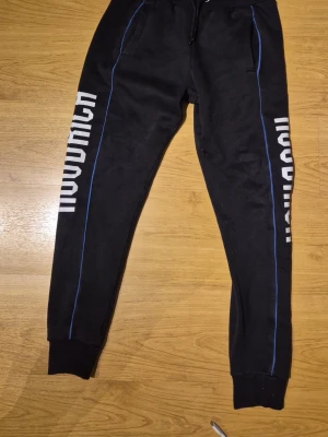 Svarta mjukisbyxor från Hoodrich XS - Svarta mjukisbyxor från Hoodrich med vit logga längs båda benen och tunna blå linjer på sidorna. Byxorna har resår i midjan med dragsko, muddar vid bensluten och två fickor fram. Perfekta för en chill och street-inspirerad look.