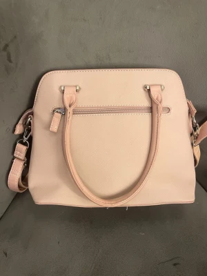 Ljusrosa handväska med axelrem - Snygg ljusrosa handväska i konstläder med både handtag och justerbar axelrem. Väskan har en stilren, fyrkantig form och silverfärgade detaljer samt dragkedjefack både fram och bak. Perfekt för dig som gillar enkel och modern design.