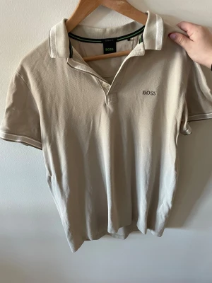 Beige pikétröja från BOSS - Klassisk beige pikétröja från BOSS med korta ärmar och krage. Tröjan har knappar framtill och diskret BOSS-logga broderad på bröstet. Perfekt till en avslappnad men stilren look.