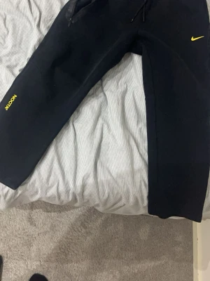Svarta NOCTA x Nike mjukisbyxor - Säljer ett par svarta mjukisbyxor från Nike och NOCTA-kollektionen. Byxorna har gul NOCTA-logga på benet och gul Nike Swoosh på höften. Utrustade med dragsko i midjan och dragkedjeförsedda fickor. Sköna och stilrena, perfekta för en chill look.