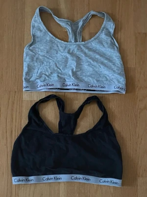 Calvin Klein bh-toppar grå & svart - Två sportiga bh-toppar från Calvin Klein, en i ljusgrått och en i svart. Båda har breda axelband och klassisk racerback-design samt elastiskt band med Calvin Klein-logga nedtill. Perfekta för träning eller chill dagar.