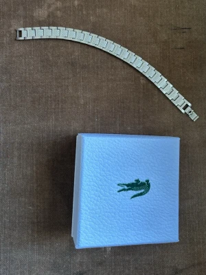 Silverfärgat armband från Lacoste - Snyggt armband i silverfärgad metall från Lacoste med rektangulära länkar och diskret Lacoste-logga på en av länkarna. Kommer i original ask med klassisk grön krokodil. Perfekt accessoar för en stilren look.