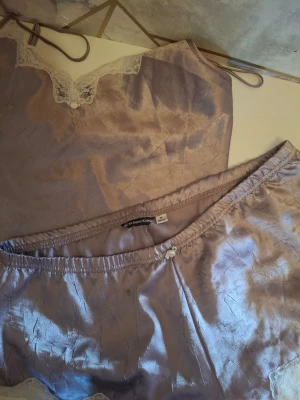 Ljuslila satinset från lindex - Sött ljuslila set i satin från KappAhl, består av ett linne med spetsdetaljer och liten rosett vid urringningen samt matchande shorts med resår i midjan och rosett. Perfekt för dig som gillar mjuka material och romantiska detaljer.