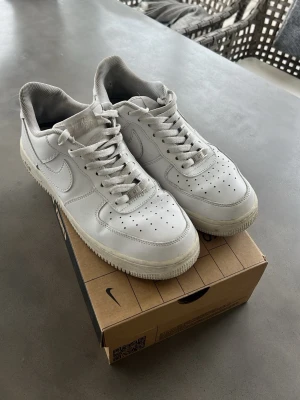 Nike Air Force 1 vita sneakers - Klassiska vita Nike Air Force 1 sneakers i lågt utförande. Skorna har en stilren design med perforerad tå, vit sula och ikoniska Swoosh-loggan på sidan. Tillverkade i läder med textilfoder och snörning framtill. Perfekta för en clean och tidlös look.